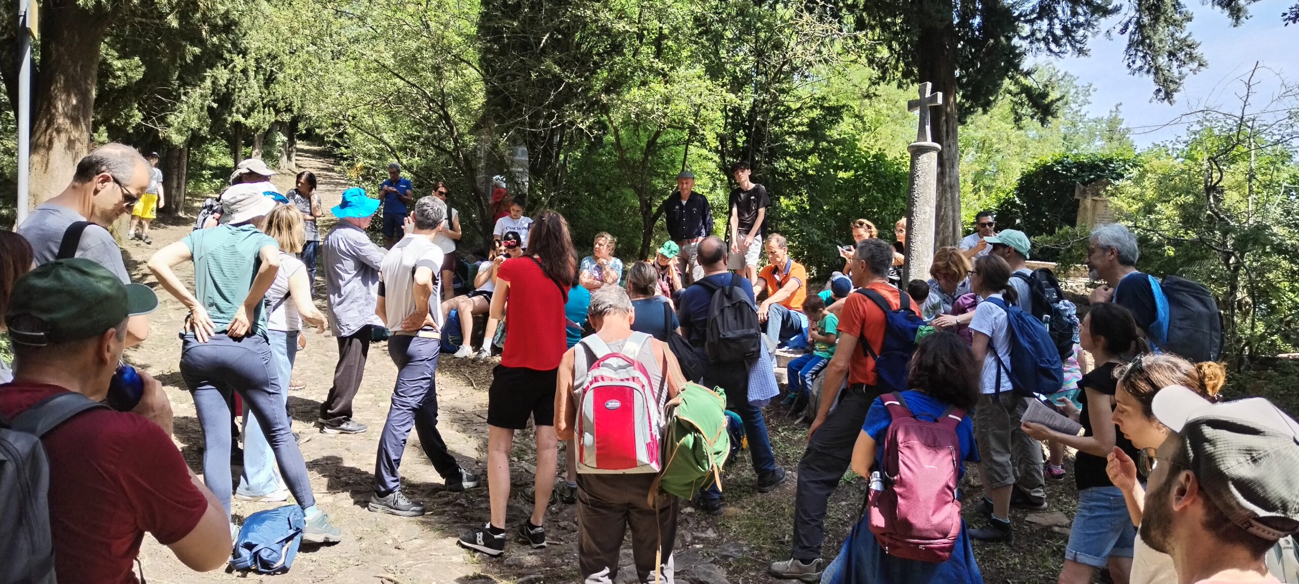 Un gruppo di famiglie con gli zaini, radunato all'ombra degli alberi attorno a una croce di pietra